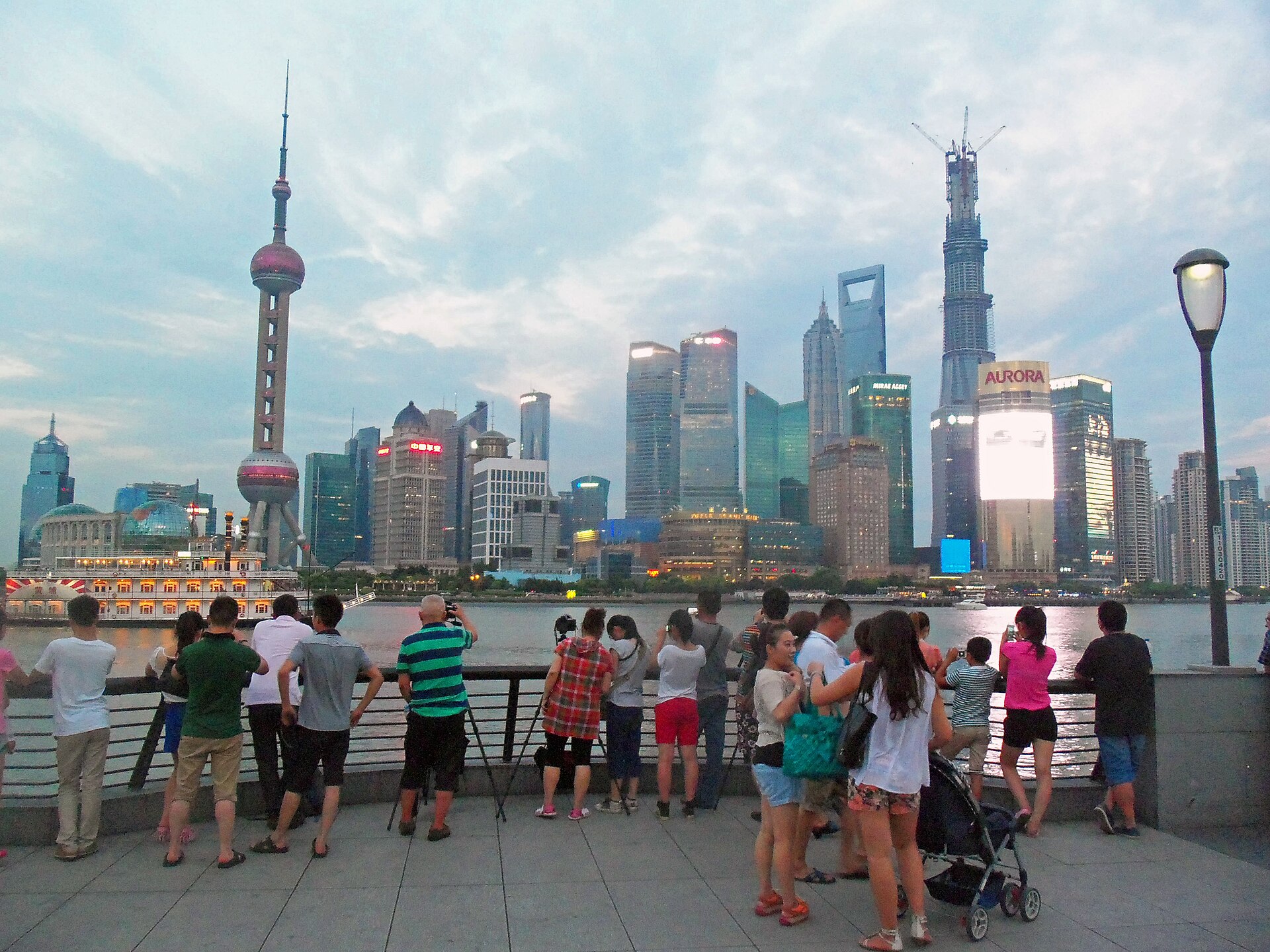 Visitors in shorts and summer shirts line the Bund promenade at dusk, photographing the Lujiazui skyline across the Huangpu River