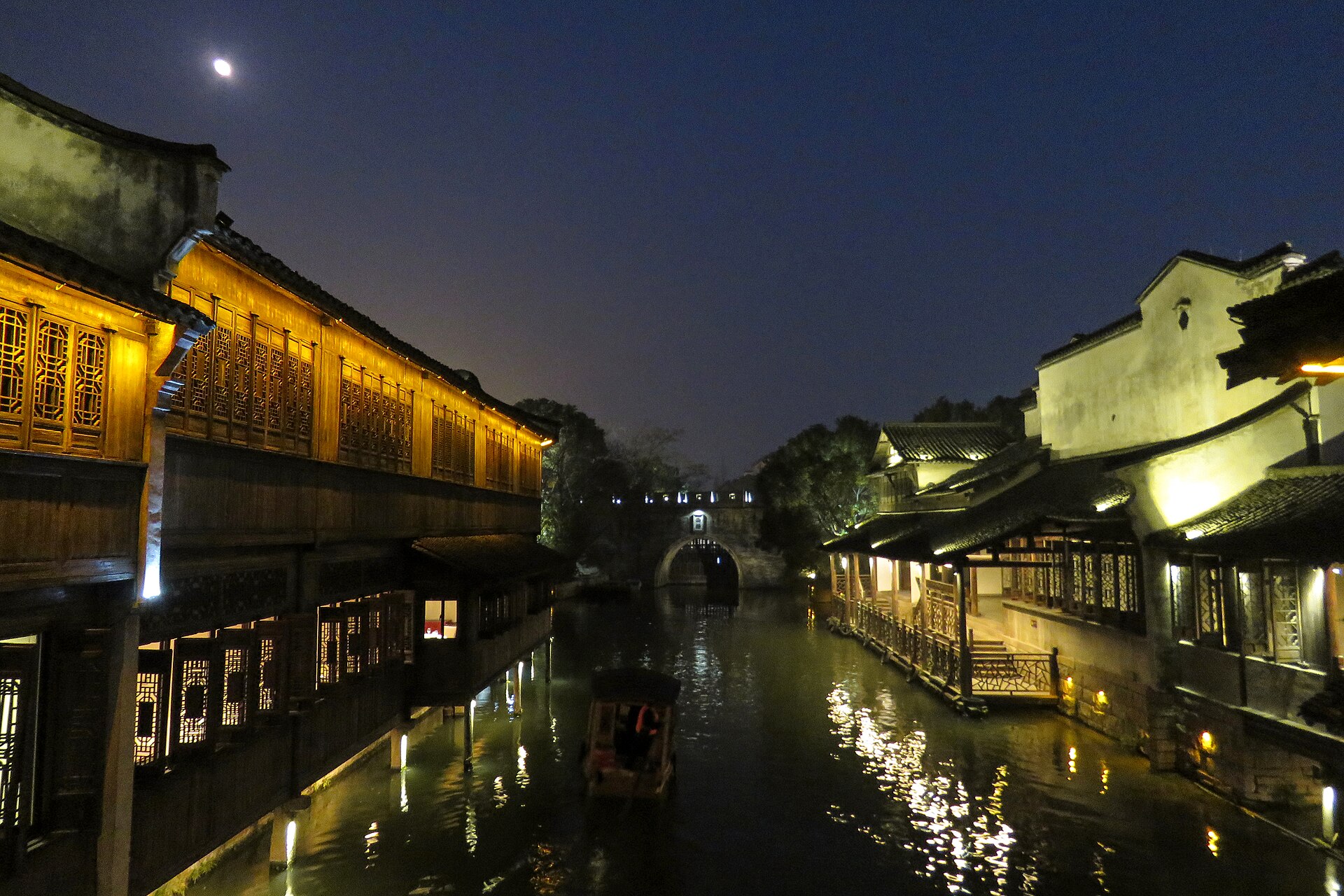 Xizha, Wuzhen, after dark — lamplit wooden lattice houses along the canal, a small boat mid-stream and an arched bridge ahead