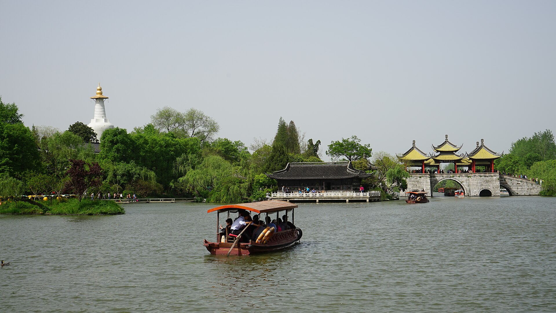 A covered boat on Slender West Lake, Yangzhou, with the White Pagoda on the left shore and the Five Pavilion Bridge ahead