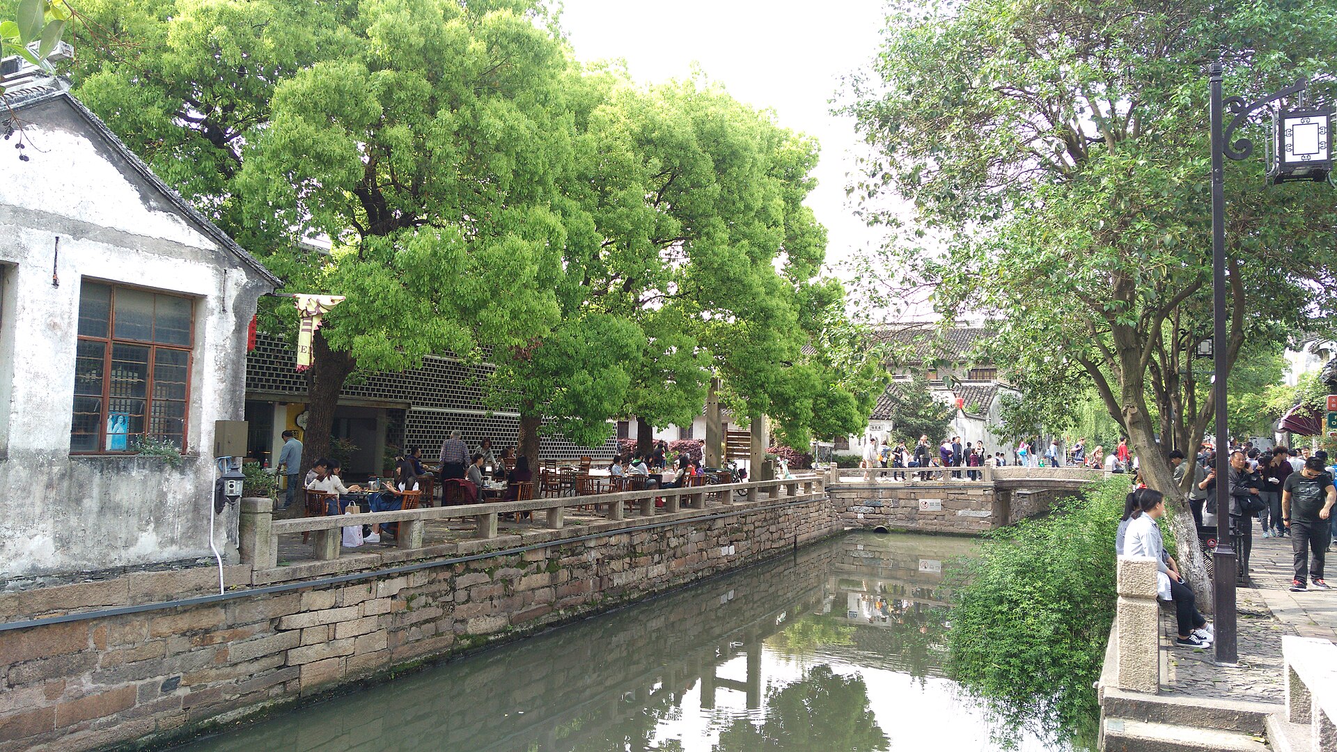 The Pingjiang canal in Suzhou, with cafe tables under camphor trees along the stone bank and a small bridge ahead